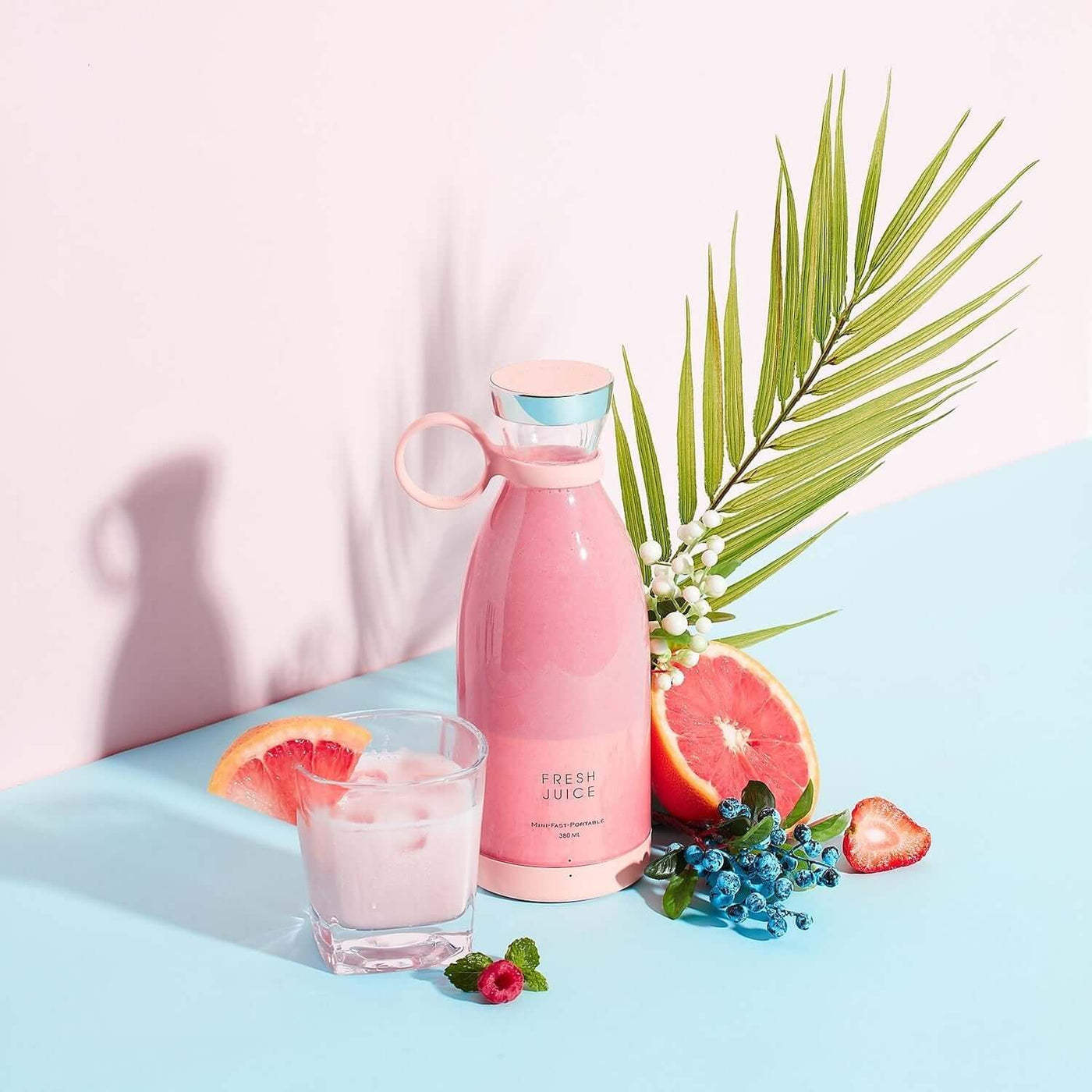 Pink Blender Bottle next to a glass of fresh juice, surrounded by fruits and greenery on a blue background.