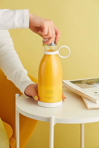 A hand holding a yellow Blender Bottle filled with fresh juice, placed on a side table with a book.
