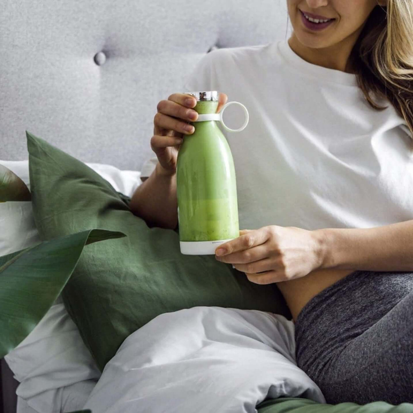 Woman holding a green Blender Bottle filled with smoothie while sitting on a bed surrounded by pillows.