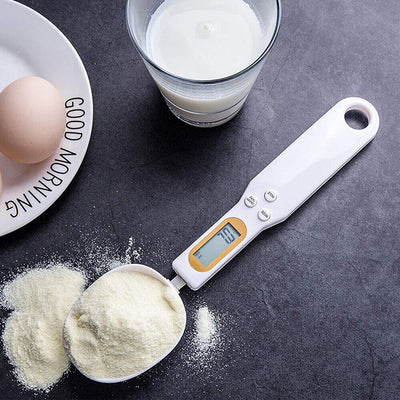 LCD digital kitchen scale spoon with backlit display, measuring ingredients like flour beside egg and milk on a black countertop.
